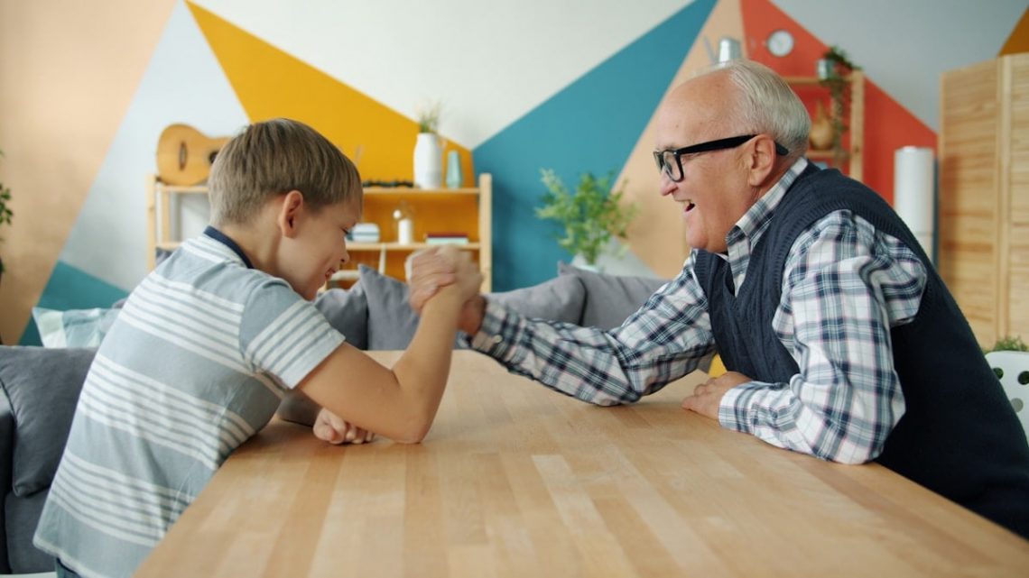 Happy child is enjoying arm wrestling with loving grandfather in apartment, people are exercising and boy is winning. Family sports and lifestyle concept.