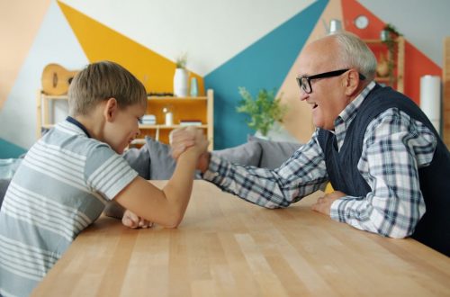 Happy child is enjoying arm wrestling with loving grandfather in apartment, people are exercising and boy is winning. Family sports and lifestyle concept.