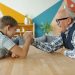 Happy child is enjoying arm wrestling with loving grandfather in apartment, people are exercising and boy is winning. Family sports and lifestyle concept.